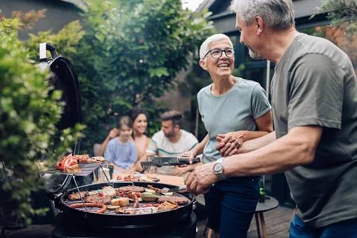 An older couple grilling on their back porch with their adult children relaxing in the backyard with the grandkids