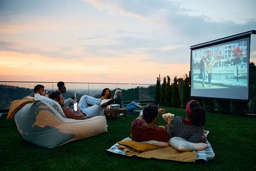 A group of friends enjoying an outdoor movie in their backyard at sunset with blankets and bag chairs spread out while the movie plays on a projector